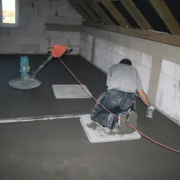 Pracownik na kolanach wygładza świeżo wylaną, ciemnoszarą posadzkę w surowym wnętrzu, obok stoi maszyna do zacierania, widoczne płyty pod kolana i poziomica.