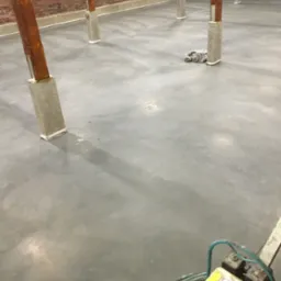 Szara, gładka posadzka w hali z drewnianymi słupami, częściowo wypolerowana, widoczna maszyna do polerowania w prawym dolnym rogu.