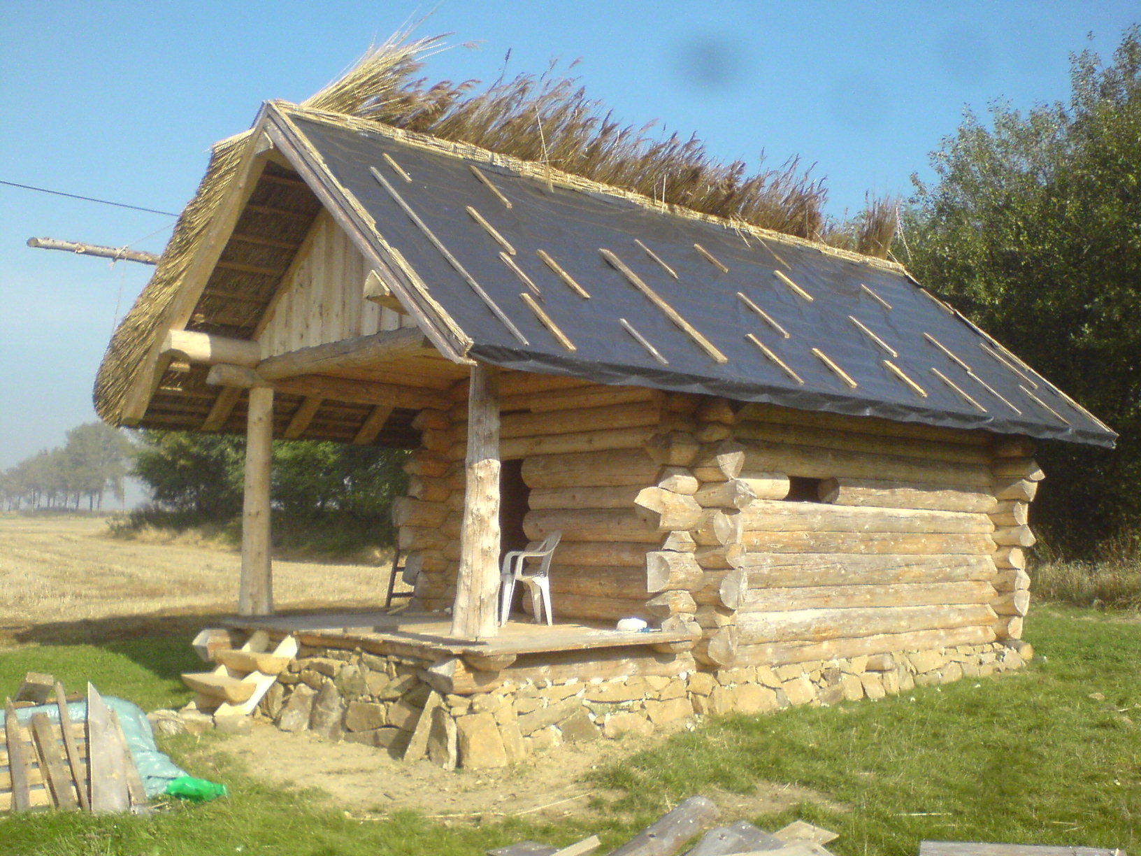 Mała drewniana chatka z bali, z kamienną podmurówką i strzechą częściowo zakrytą czarną folią, w tle pole i drzewa.