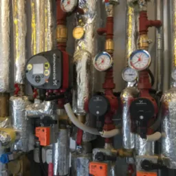 Złożona instalacja grzewcza z pompami obiegowymi Grundfos, manometrami i zaworami, rury zaizolowane folią aluminiową, widoczne elementy automatyki Belimo.