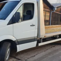 Biały samochód dostawczy z platformą załadunkową przewożący drewniane deski, zaparkowany na betonowym placu przed drewnianym budynkiem.