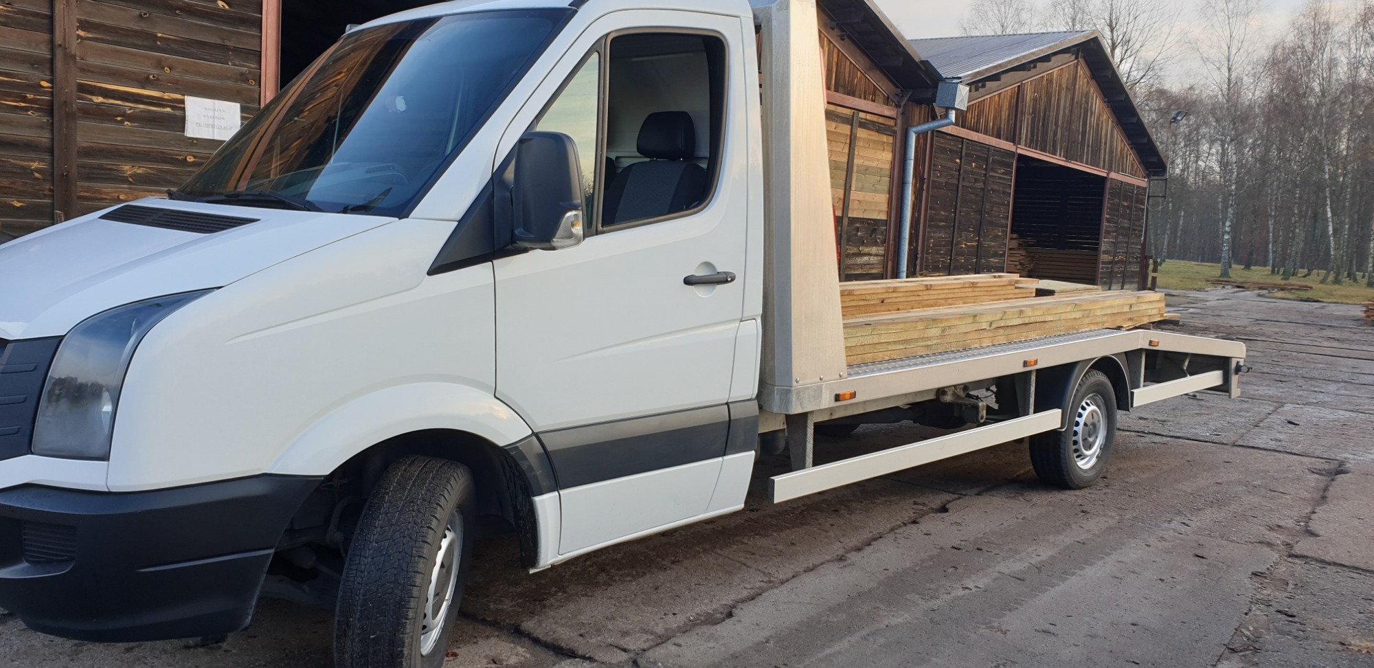 Biały samochód dostawczy z platformą załadunkową przewożący drewniane deski, zaparkowany na betonowym placu przed drewnianym budynkiem.