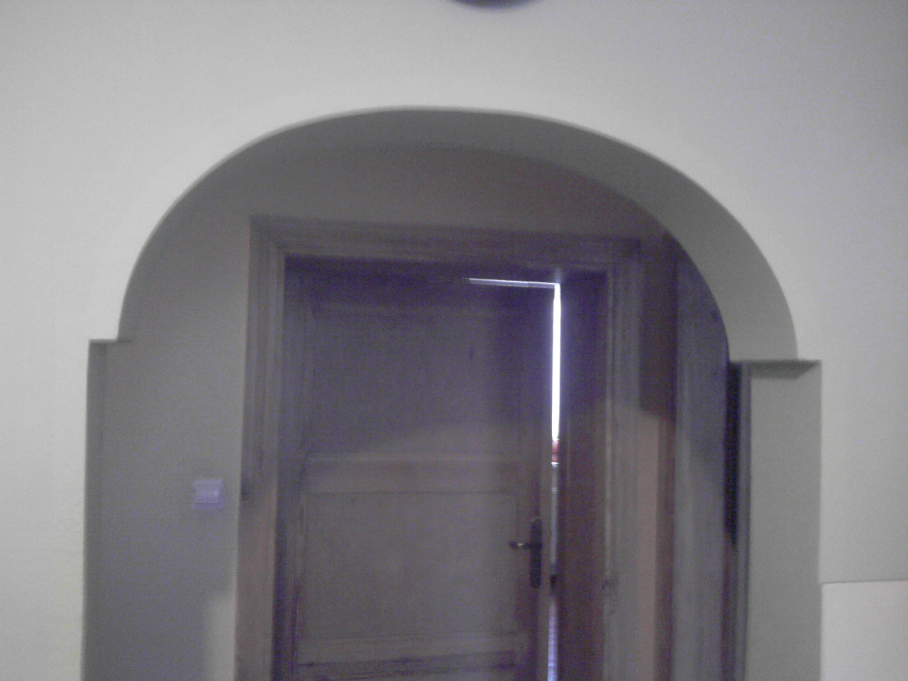 Otwarty drewniany portal drzwiowy z łukowatym wykończeniem, świeżo pomalowany na szaro, widoczny fragment ściany z włącznikiem.