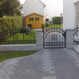 Czarna, metalowa brama z ozdobnymi detalami, osadzona na kamiennych słupkach, prowadząca do ogrodu z zielonym trawnikiem i murowaną ścieżką.