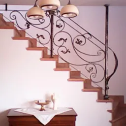 Kute, zdobione balustrady schodowe w brązowej tonacji, z dekoracyjnym oświetleniem. Na dole komody z ozdobami.