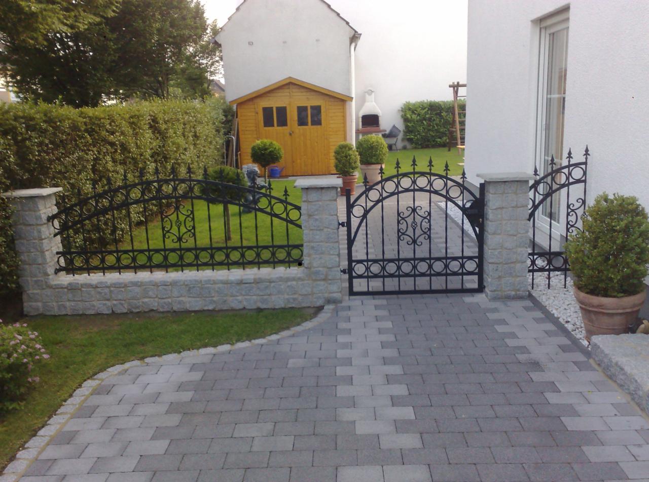 Czarna, metalowa brama z ozdobnymi detalami, osadzona na kamiennych słupkach, prowadząca do ogrodu z zielonym trawnikiem i murowaną ścieżką.