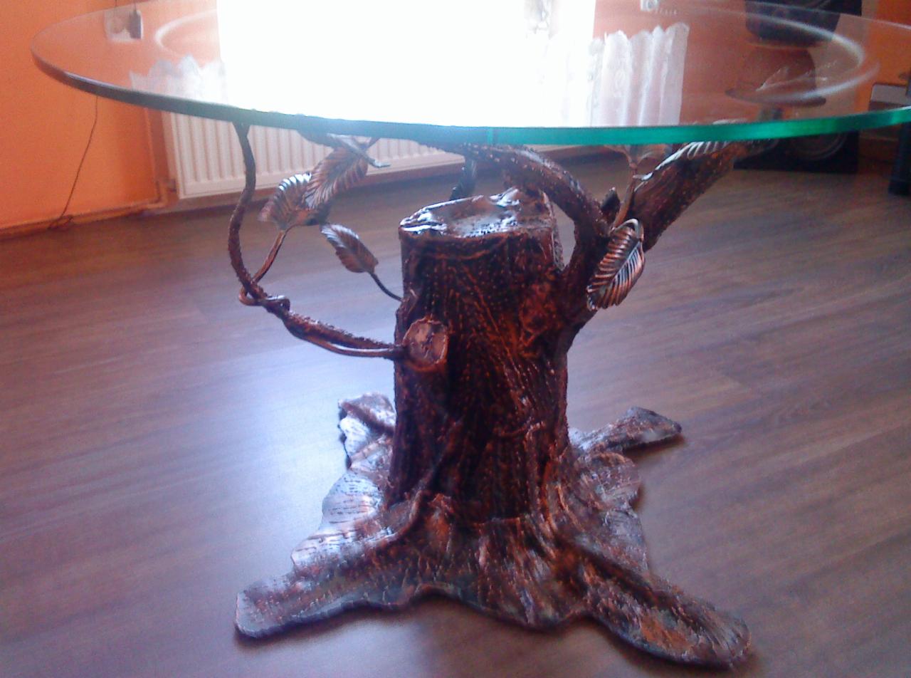 Stół z blatem ze szkła i podstawą imitującą pień drzewa z metalowymi liśćmi, na drewnianej podłodze, oświetlony naturalnym światłem.