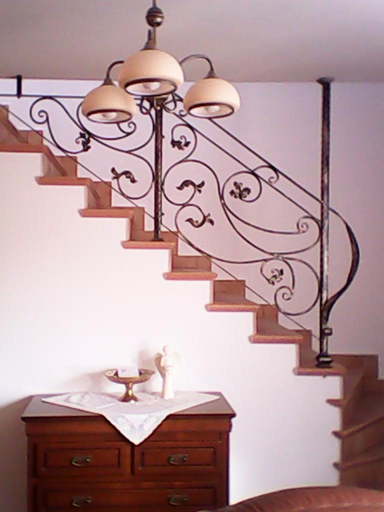 Kute, zdobione balustrady schodowe w brązowej tonacji, z dekoracyjnym oświetleniem. Na dole komody z ozdobami.