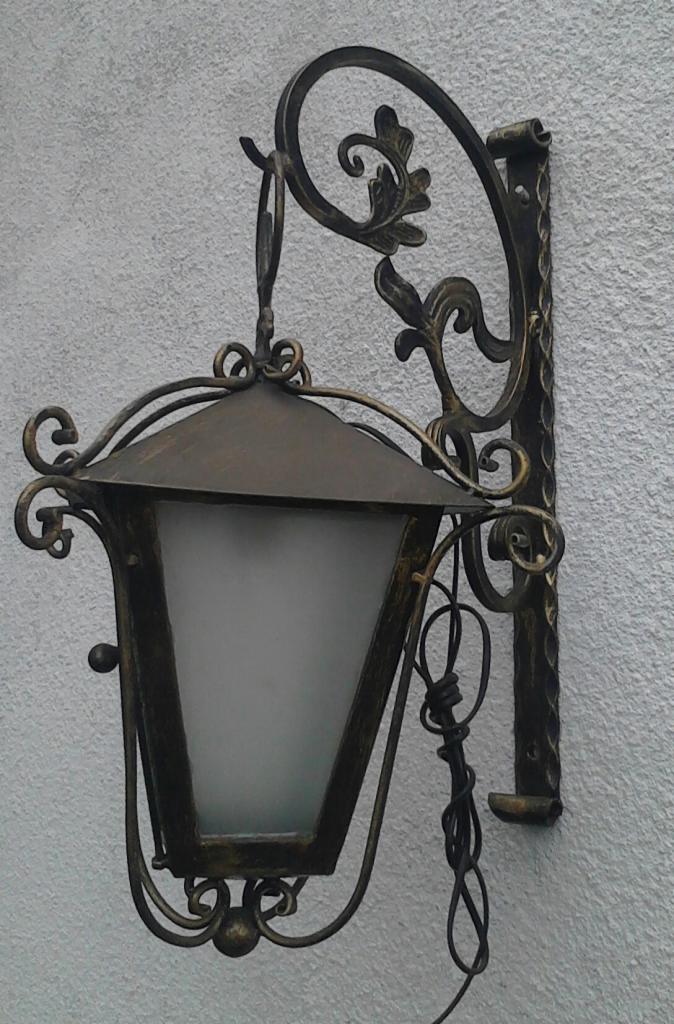 Kuta, ozdobna lampa ścienna w stylu retro, z dekoracyjnymi elementami roślinnymi i szklanym kloszem, zamontowana na jasnej, teksturowanej ścianie.