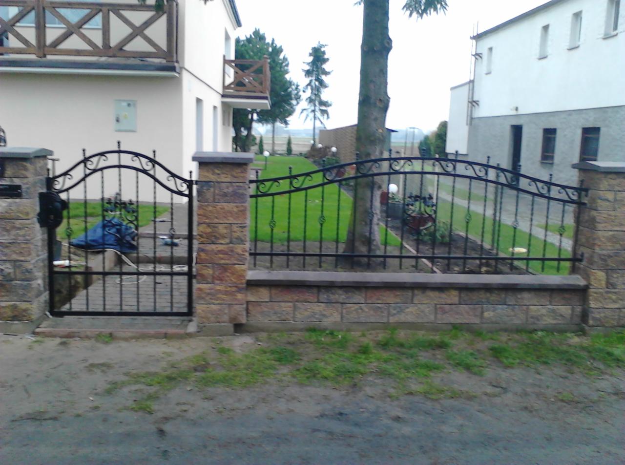 Kuta brama i ogrodzenie z kamiennymi słupkami na tle domu z balkonem i zielonego trawnika. Delikatne zdobienia metalowe. Widok z zewnątrz posesji.