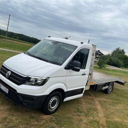 Przedsiębiorstwo Usługowo-Handlowe Sigma Dariusz Nowatkiewicz - Biała laweta VW Crafter na trawiastym terenie, widok z przodu pod kątem. Antena na dachu, tablica rejestracyjna widoczna. Krajobraz wiejski w tle, pochmurne niebo.