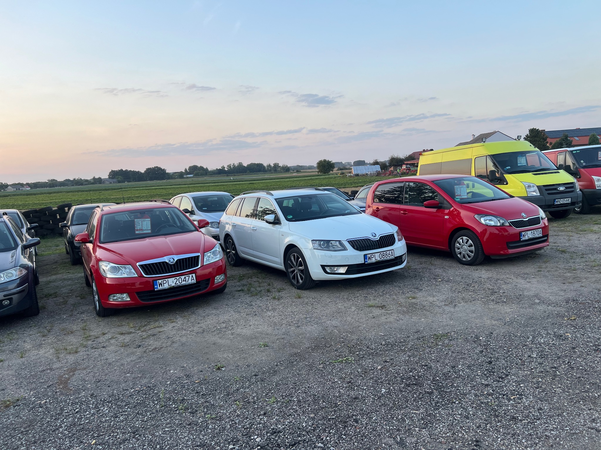 Flota pojazdów Skoda w Małej Wsi, gotowa do transportu. Widoczne auta osobowe i dostawcze, ustawione na żwirowym placu, w tle pole uprawne i zachodzące słońce.