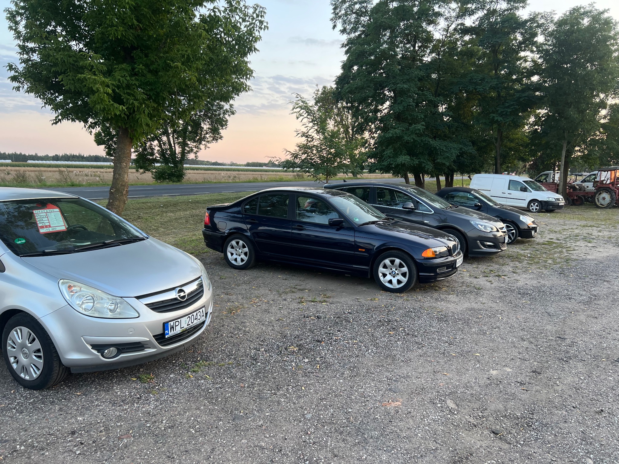 Rząd zaparkowanych samochodów różnych marek i modeli na żwirowym parkingu, w tle drzewa i pole uprawne, widoczne tablice rejestracyjne.