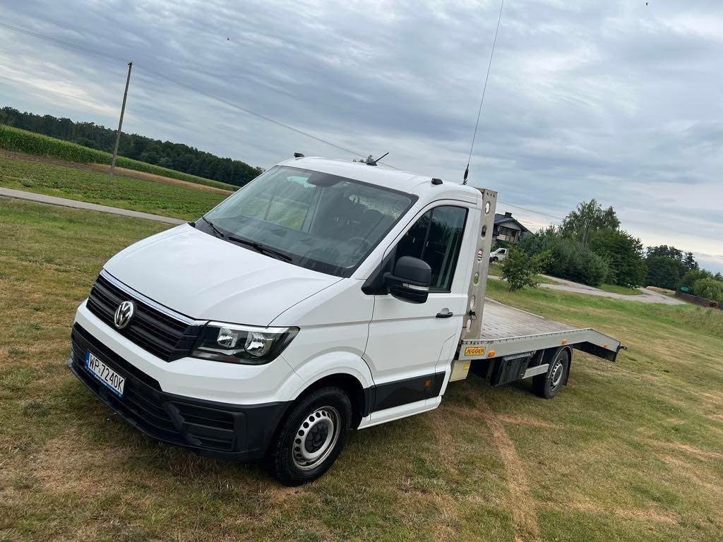 Biała laweta VW Crafter na trawiastym terenie, widok z przodu pod kątem. Antena na dachu, tablica rejestracyjna widoczna. Krajobraz wiejski w tle, pochmurne niebo.