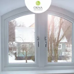 Białe, dwuskrzydłowe okno z łukowym górnym panelem, widok na zasypany śniegiem ogród z domem w tle, delikatne światło słoneczne.