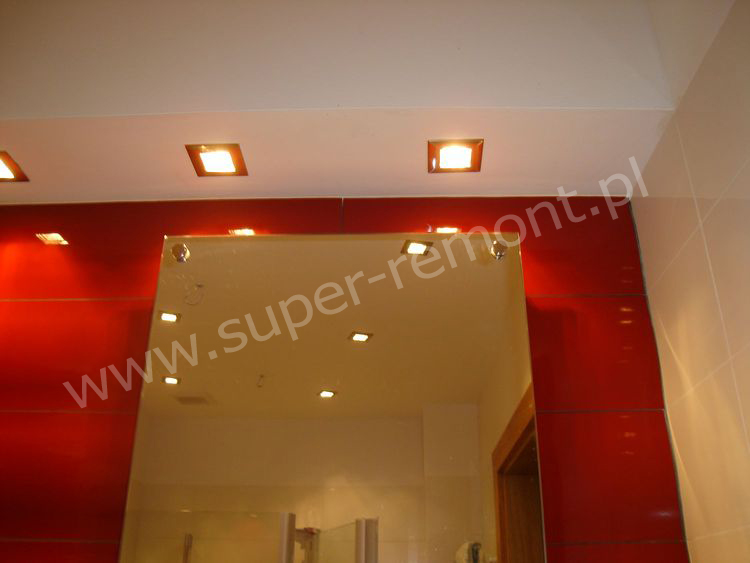 Czerwona ściana z lustrem i kwadratowymi lampkami sufitowymi w łazience w Warszawie, widoczne odbicie wnętrza.