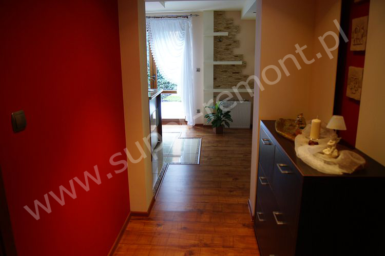 Wnętrze przedpokoju z czerwoną ścianą, drewnianą podłogą i komodą z dekoracjami, widok na pokój z oknem i kamienną dekoracją ścienną.