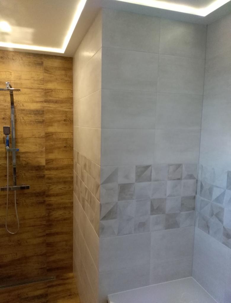 Wykończona łazienka z kabiną prysznicową: ściany wyłożone jasnymi płytkami z geometrycznym wzorem i panelami imitującymi drewno, oświetlenie sufitowe LED.