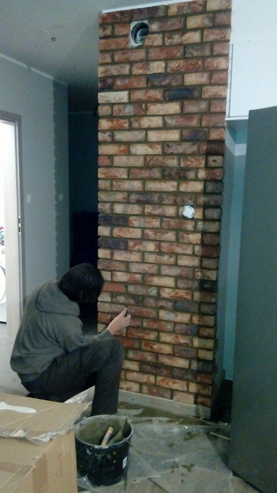 Montaż dekoracyjnej ściany z cegły wewnątrz pomieszczenia. Widoczny murarz w trakcie pracy, narzędzia i fragment ściany z otworem wentylacyjnym.