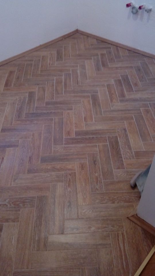 Ukończona podłoga z płytek imitujących drewno, ułożonych w jodełkę, z widocznymi listwami przypodłogowymi i fragmentem ściany. Ciepłe odcienie brązu.