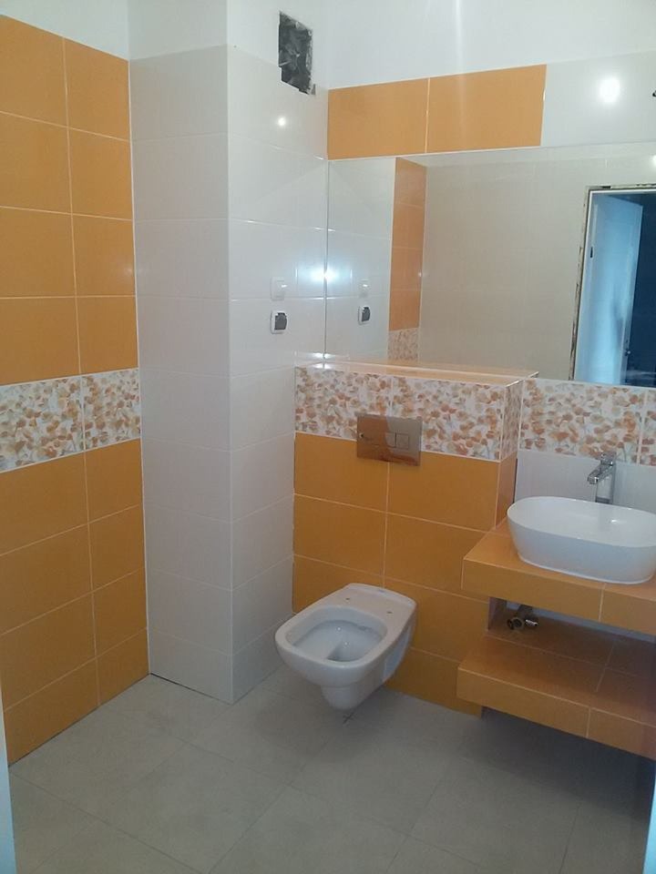 Wykończona łazienka z pomarańczowymi i białymi płytkami, umywalką nablatową i podwieszaną toaletą. Widoczne lustro i fragment otwartych drzwi.