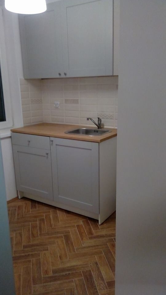 Jasna kuchnia po remoncie: szare szafki z drewnianym blatem, białe płytki na ścianie i podłoga ułożona w jodełkę. Widok na umywalkę i baterię.