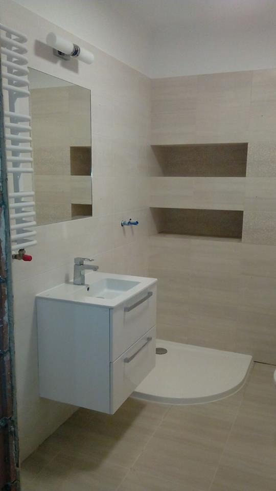 Wykończona łazienka z beżowymi płytkami, wiszącą szafką z umywalką, lustrem i grzejnikiem drabinkowym. Widoczne wnęki w ścianie i brodzik.