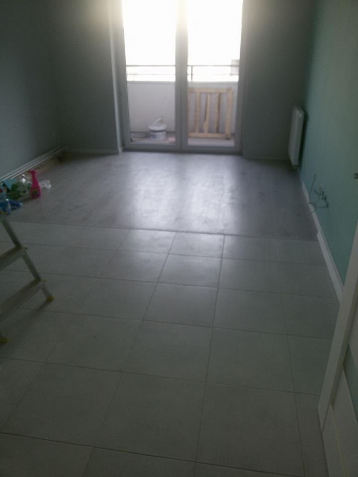 Wnętrze pokoju z nową, jasną podłogą z płytek. Widoczne drzwi balkonowe z barierką. Część podłogi jeszcze nie wykończona. Na podłodze leżą narzędzia i materiały.