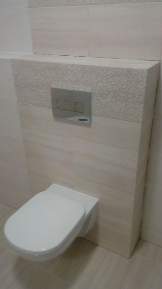 Podwieszana toaleta z beżowymi płytkami imitującymi drewno, dekoracyjny wzór na zabudowie stelaża, chromowany przycisk spłukujący. Minimalistyczny design.