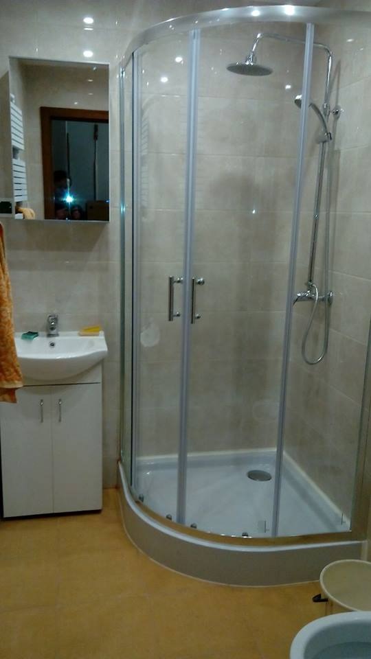 Wykończona łazienka z kabiną prysznicową narożną, umywalką z szafką i lustrem. Beżowe płytki na ścianach, jasna podłoga. Widoczne detale wykończeniowe.