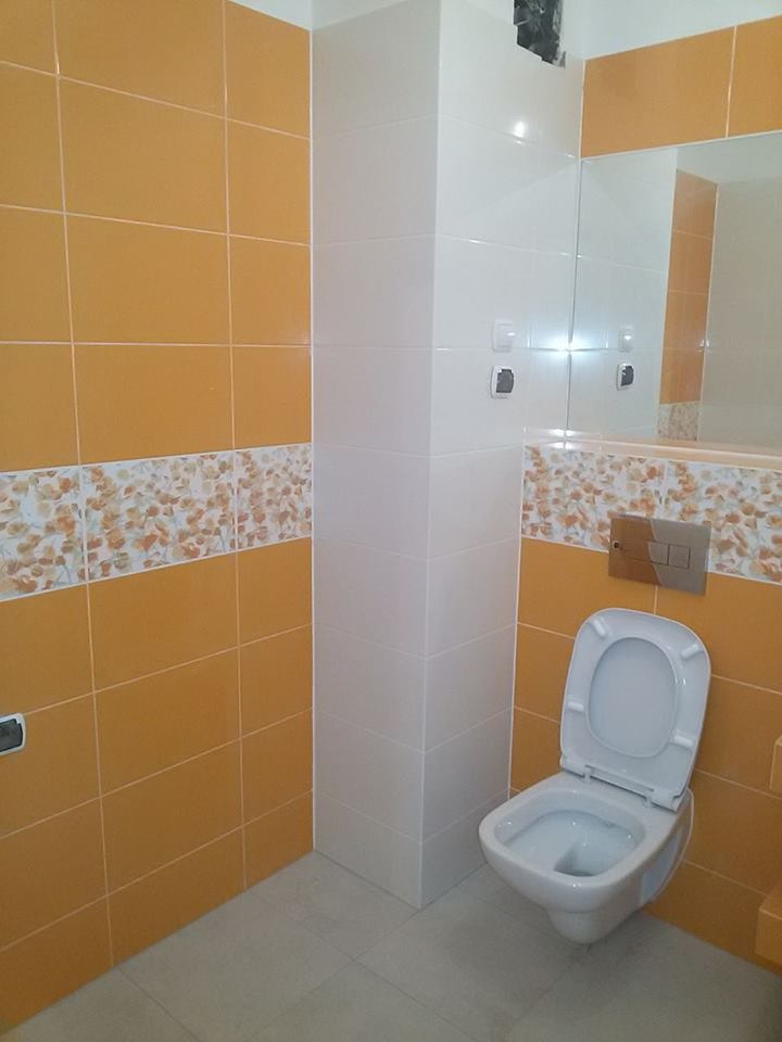 Wykończona łazienka z pomarańczowymi i białymi płytkami, dekoracyjnym pasem z motywem kwiatowym i podwieszaną toaletą. Widoczne elementy armatury i lustro.