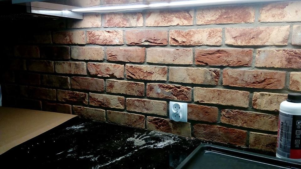 Ściana z cegły dekoracyjnej w kuchni, z widocznym gniazdkiem elektrycznym i fragmentem blatu. Oświetlenie LED podkreśla fakturę cegieł.