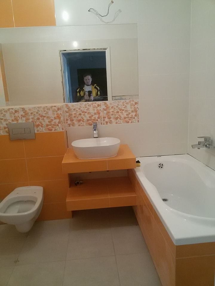 Wykończona łazienka z pomarańczowymi akcentami: płytki, szafka pod umywalką i obudowa wanny. Widoczne białe sanitariaty i lustro z odbiciem osoby.