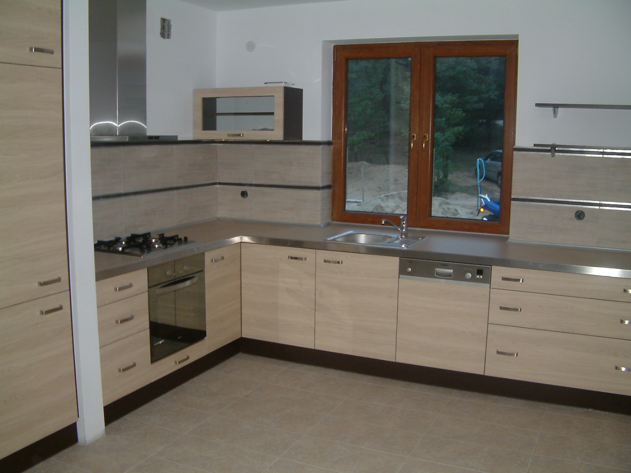 Jasna kuchnia z drewnianymi frontami, stalowym blatem i oknem z widokiem na zieleń, w tle widoczny samochód i plac budowy.