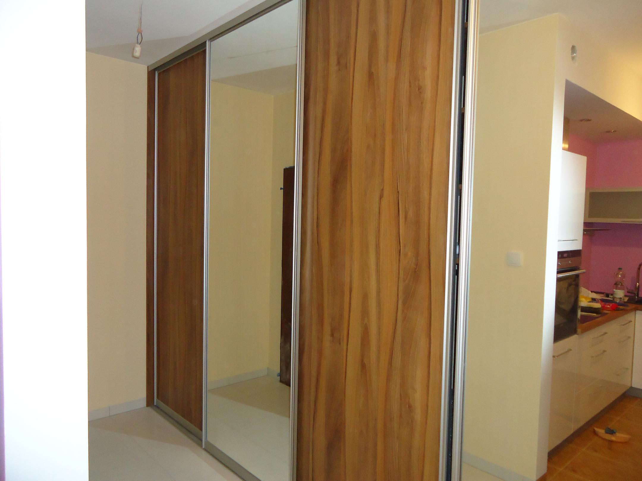 Trzydrzwiowa szafa przesuwna z lustrzanymi i drewnianymi frontami w jasnym wnętrzu, widoczna kuchnia w tle.