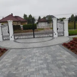 Otwarta, kuta brama wjazdowa z białymi, kolumnowymi słupkami, automatyką i dekoracyjnym wzorem, prowadząca do posesji z kostki brukowej, w tle widoczny dom z czerwonym dachem.
