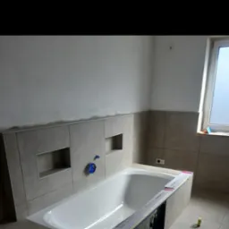 Biała wanna w trakcie montażu w łazience wyłożonej beżowymi płytkami z wnękami, widoczne narzędzia i materiały budowlane, ściana częściowo nieotynkowana, okno z matową szybą.