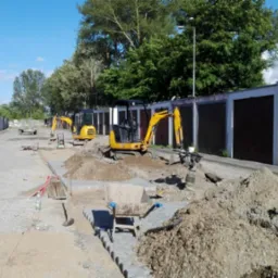 Dwa żółte minikoparki przygotowują teren pod nową nawierzchnię wzdłuż rzędu garaży; widoczny fragment ułożonej kostki i hałdy ziemi.