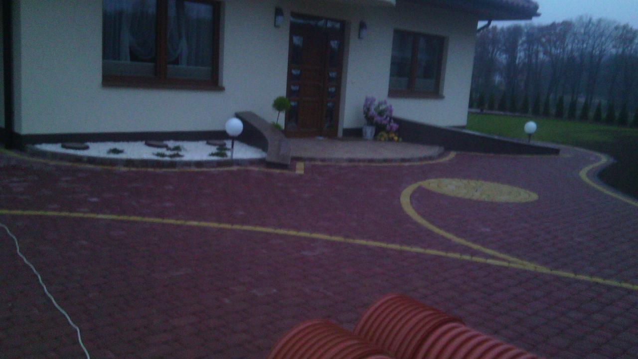 Ogród z kostki brukowej w odcieniach czerwieni i żółci, z elementami dekoracyjnymi: białym żwirem, roślinami i oświetleniem, na tle domu z brązowymi oknami.