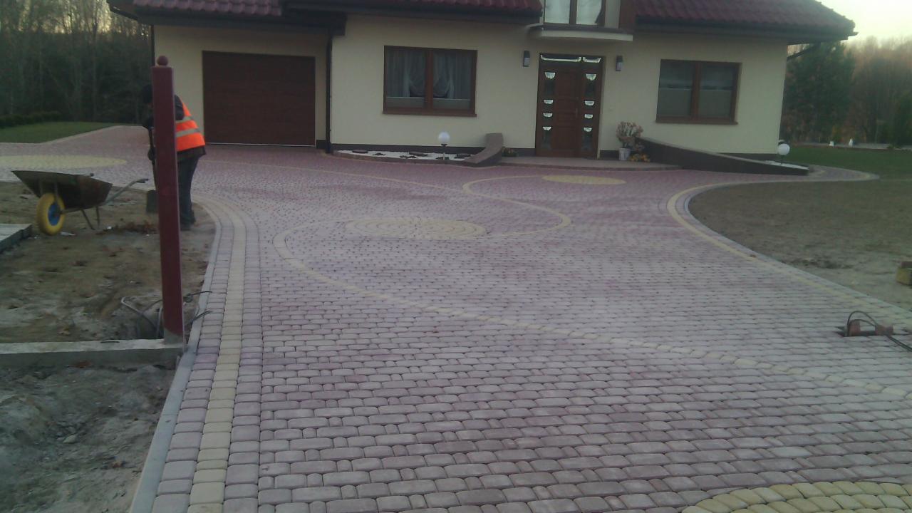 Ułożona kostka brukowa w odcieniach szarości i beżu tworząca wzór na podjeździe przed domem z brązową stolarką. Widoczny fragment trawnika i prace brukarskie.