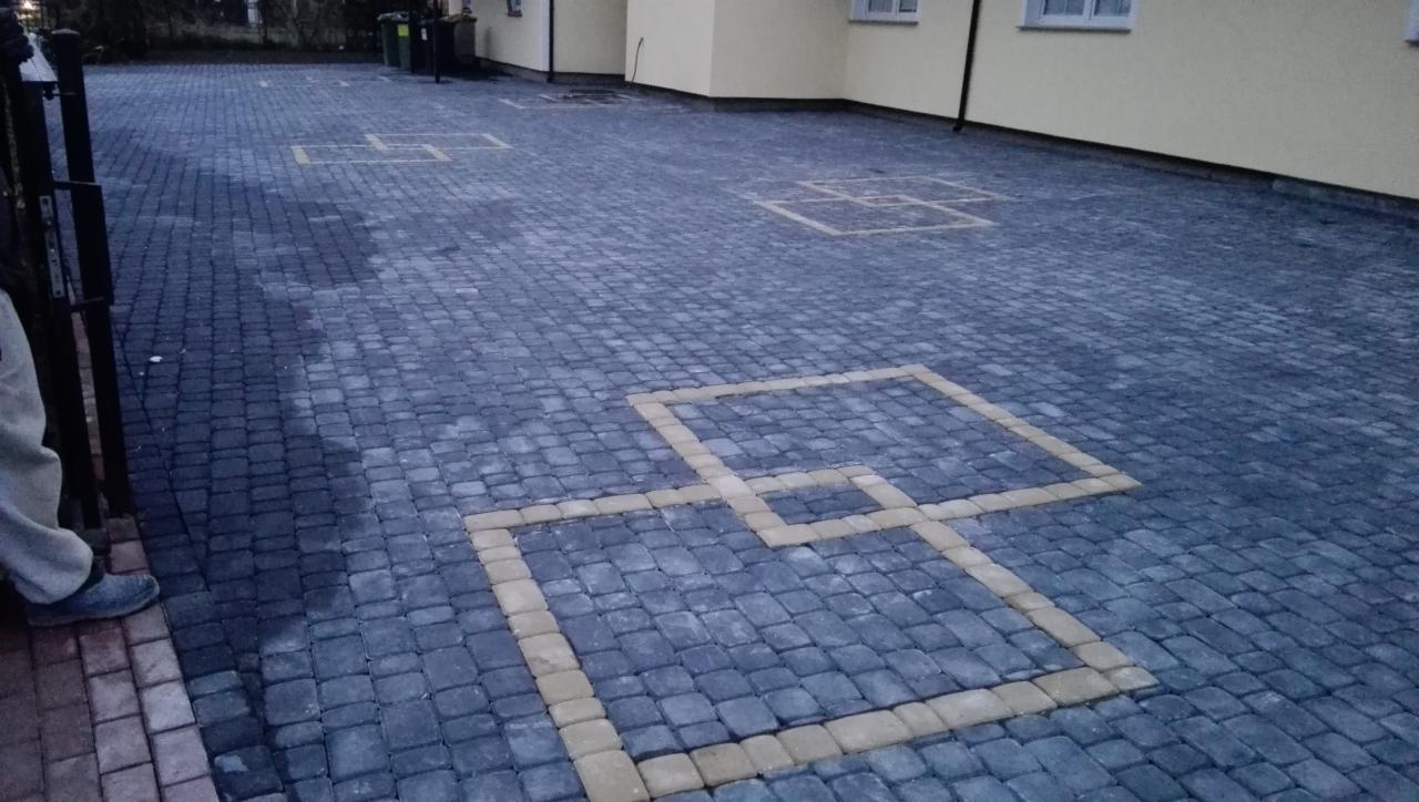 Wykończony plac z szarej kostki brukowej z żółtymi obramowaniami kwadratów, widoczne fragmenty budynku i ogrodzenia.