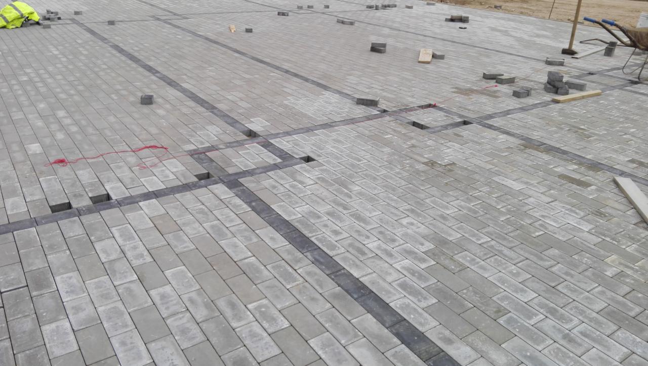 Kładzenie szarej kostki brukowej z ciemniejszymi obramowaniami, widoczne braki w ułożeniu i narzędzia pracy, w tle żółta kamizelka odblaskowa.