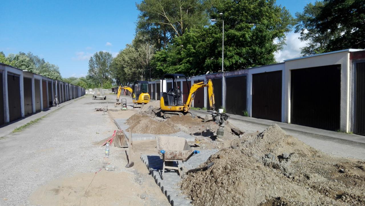 Dwa żółte minikoparki przygotowują teren pod nową nawierzchnię wzdłuż rzędu garaży; widoczny fragment ułożonej kostki i hałdy ziemi.