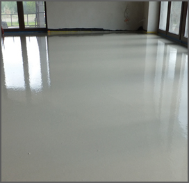 Gładka, jasna wylewka anhydrytowa na ogrzewaniu podłogowym, widoczne okna z odbiciem światła, ściany w stanie surowym.