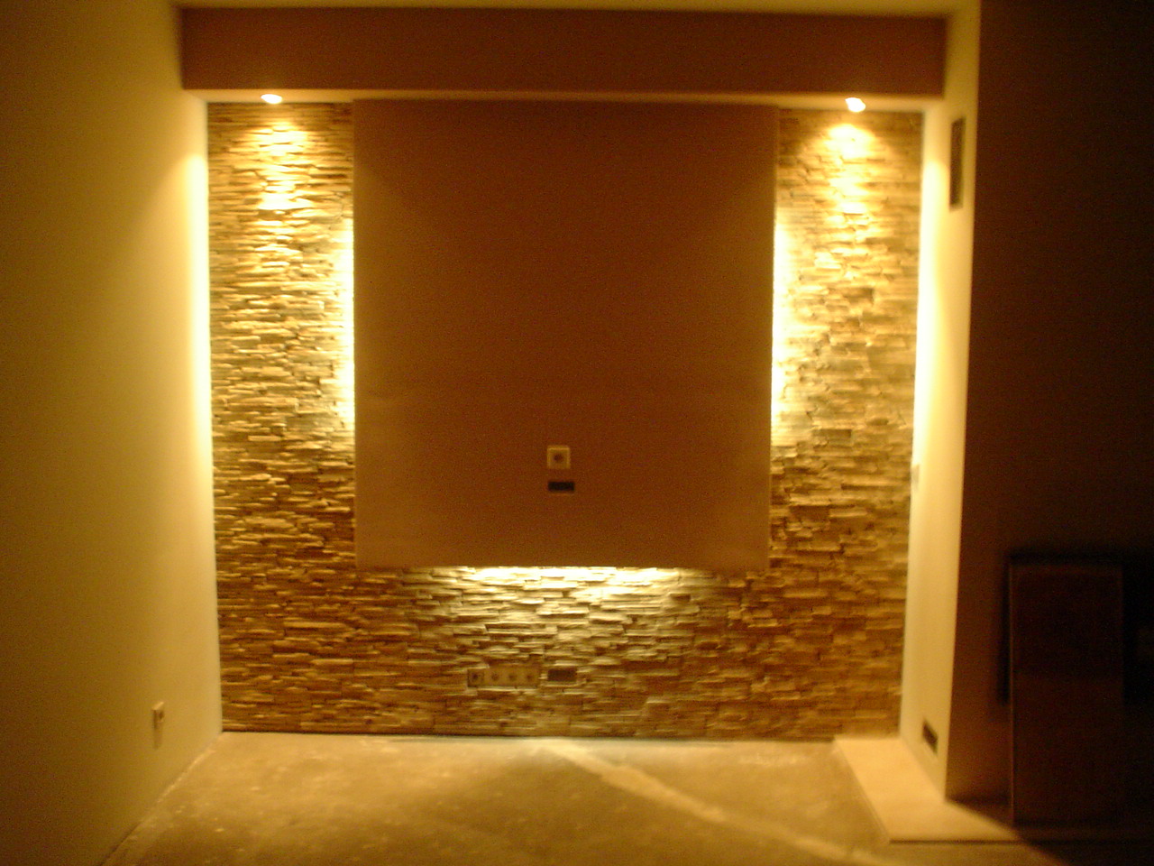 Ściana akcentująca z kamienia dekoracyjnego podświetlona punktowymi lampami w salonie, widoczne gniazdka elektryczne i pusta przestrzeń pod ścianą.