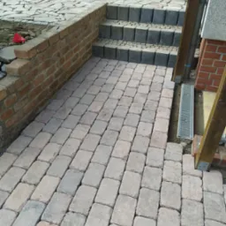 Ułożona kostka brukowa w odcieniach szarości i beżu, przylegająca do ceglanego murku i schodów z szarych kostek, z widocznym odpływem liniowym.