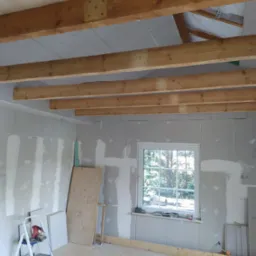 Wnętrze w trakcie montażu płyt kartonowo-gipsowych na ścianach i suficie, widoczne drewniane belki stropowe, drabina z narzędziami i okno z widokiem na zieleń.