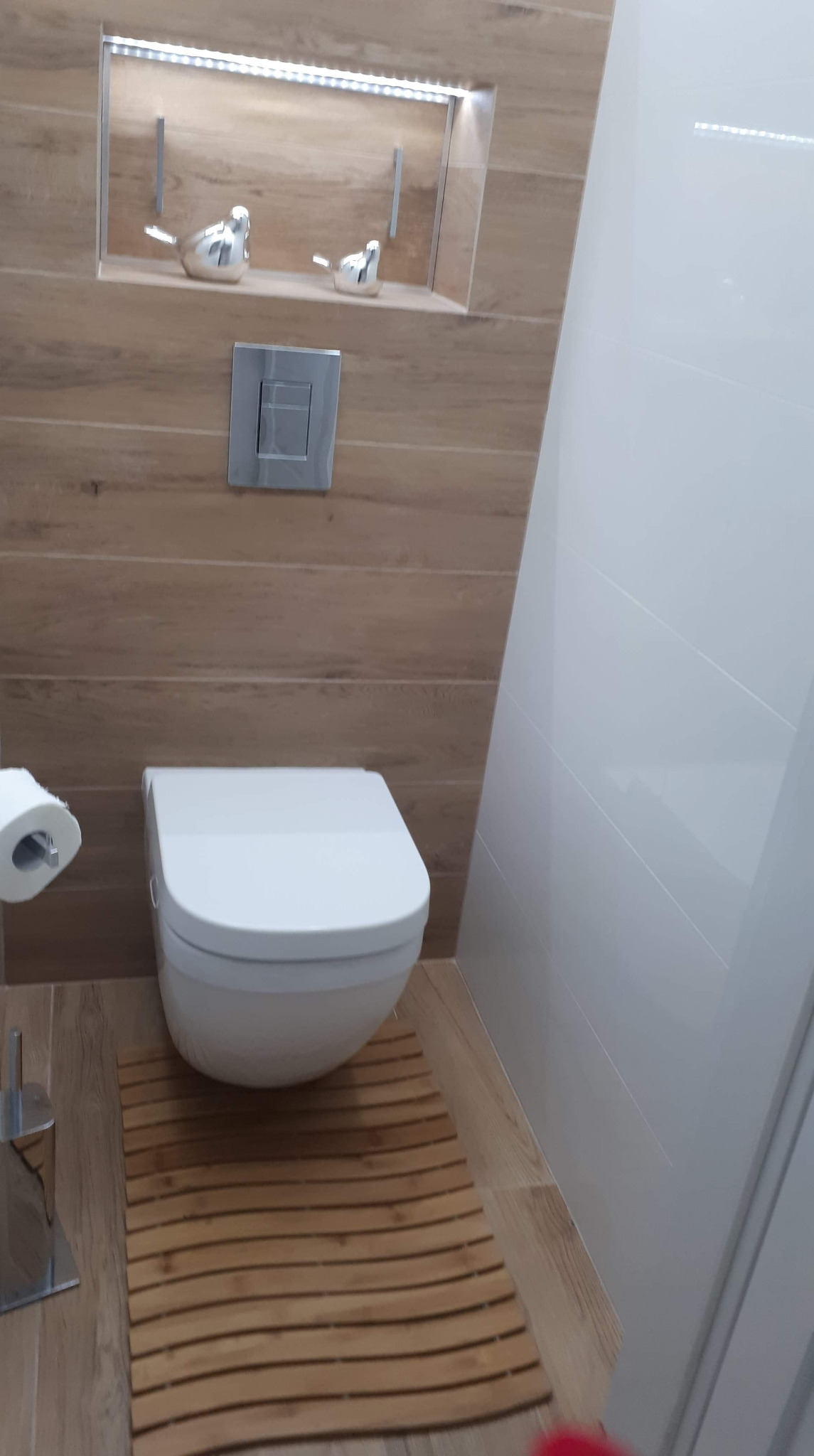 Nowoczesna toaleta podwieszana z drewnopodobnymi płytkami, chromowanym przyciskiem spłukującym i dekoracyjną wnęką z oświetleniem LED, w której umieszczono dwie srebrne figurki ptaków.