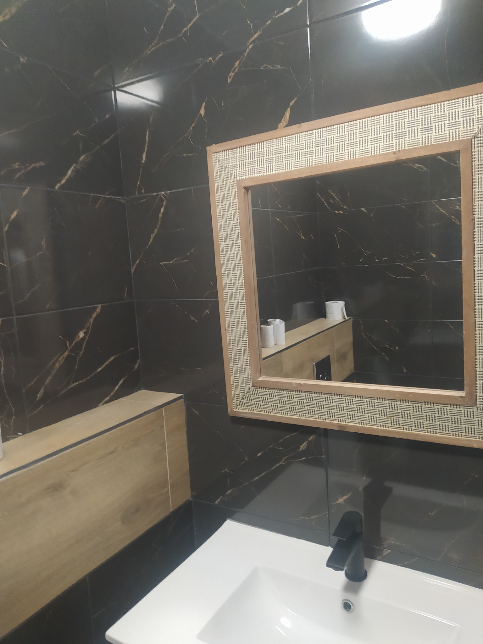 Nowoczesna łazienka z czarnymi płytkami imitującymi marmur, białą umywalką z czarną baterią i lustrem w ramie z plecionki.