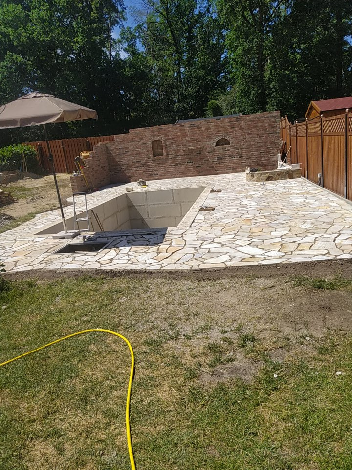 Budowa basenu ogrodowego z kamienną posadzką i ceglaną ścianą w tle, widoczna drabinka basenowa i żółty wąż ogrodowy na trawie.
